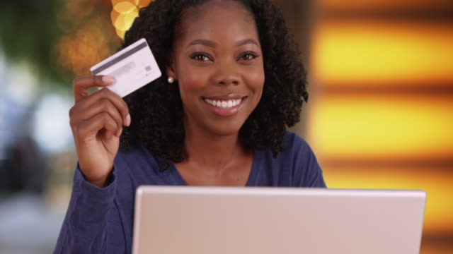 Cheerful black female in outdoor setting holding up credit card and smiling at camera. Smiling portrait of smart, financially independent young woman posing with laptop and card. 4k